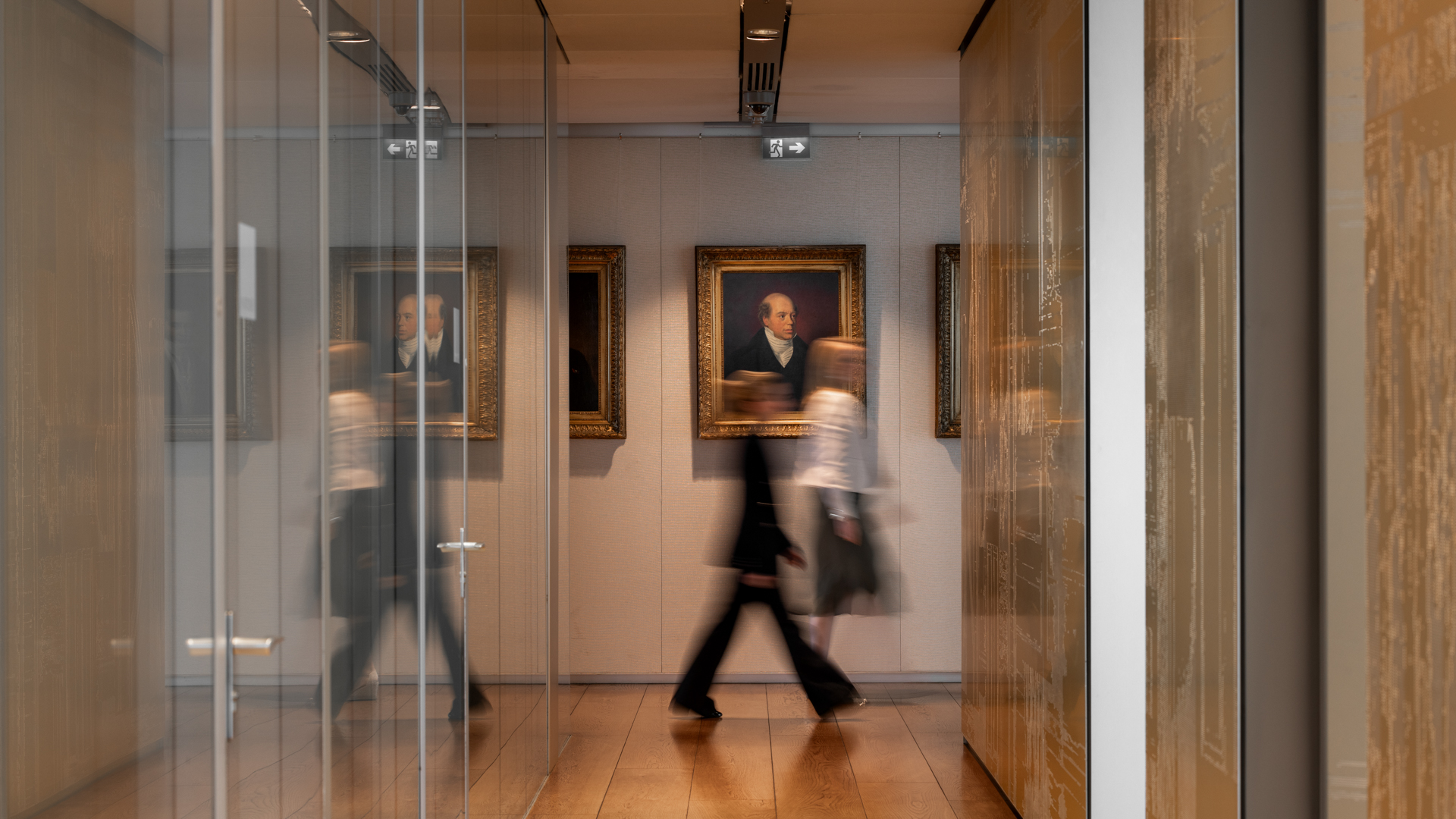 People move through a corridor lined with glass walls and historical portraits, blending modern office life with a sense of tradition.