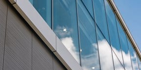Modern glass office façade reflecting clouds against a clear blue sky, highlighting the building's clean lines and contemporary design.