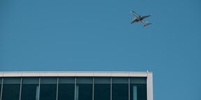 Commercial aircraft flying above a modern office building against a clear blue sky.