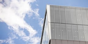 Corner of a modern glass office building set against a bright blue sky with scattered white clouds.