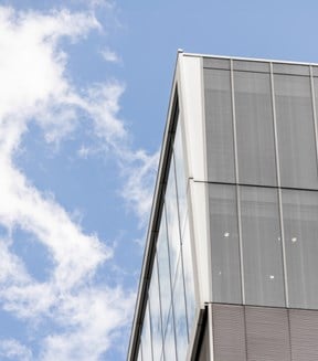 Corner of a modern glass office building set against a bright blue sky with scattered white clouds.