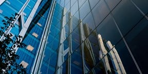Upward view of a modern glass office building with reflective façades mirroring neighbouring structures against a clear blue sky.