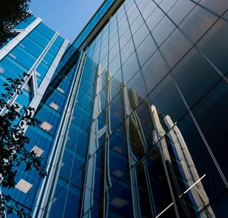 Upward view of a modern glass office building with reflective façades mirroring neighbouring structures against a clear blue sky.