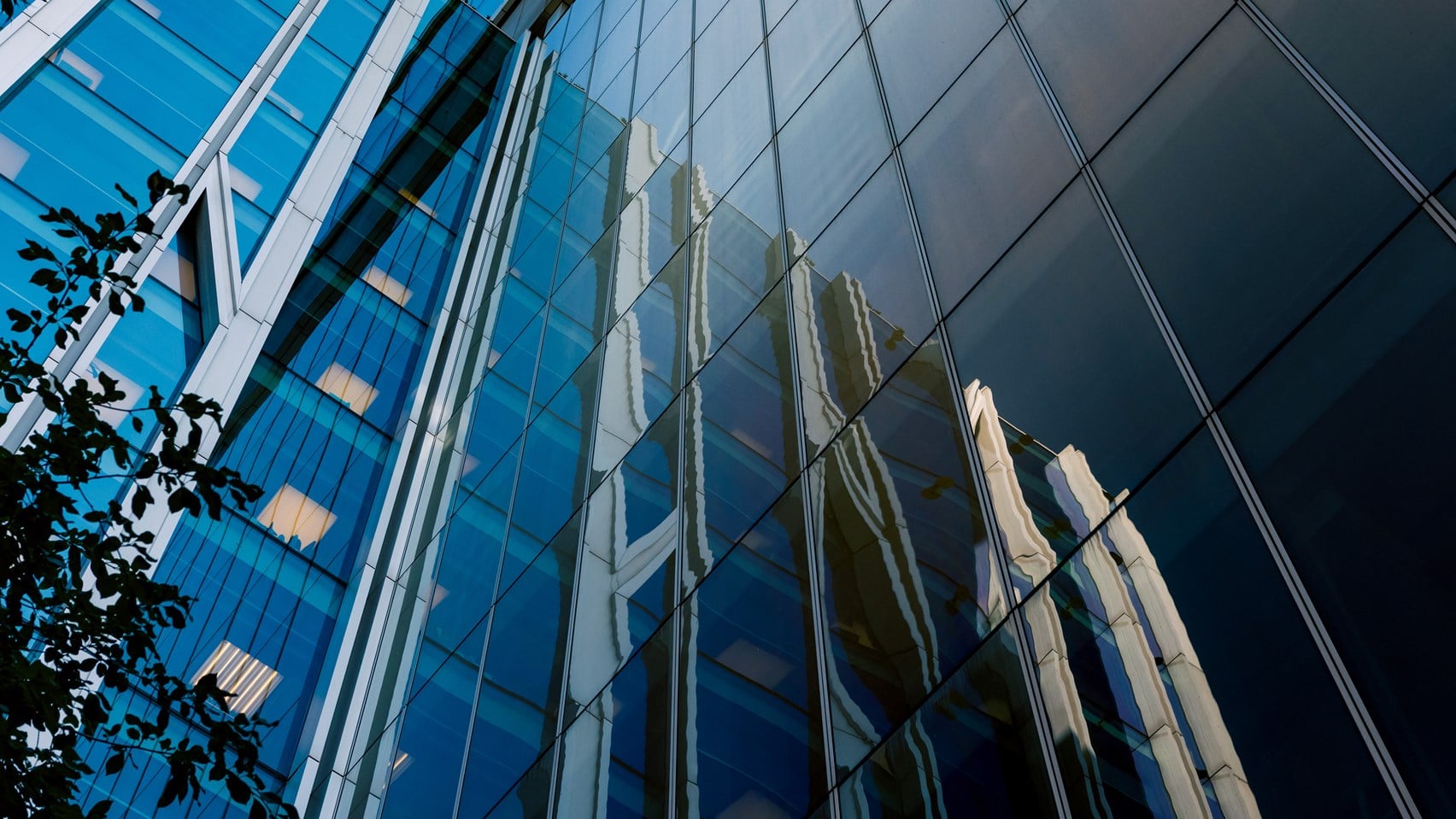Upward view of a modern glass office building with reflective façades mirroring neighbouring structures against a clear blue sky.