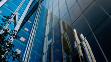 Upward view of a modern glass office building with reflective façades mirroring neighbouring structures against a clear blue sky.