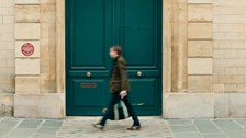 A person walks past a large teal-blue door set within a cream stone façade, captured with slight motion blur. The cobbled pavement and symmetrical architecture create a clean, minimalist street scene.