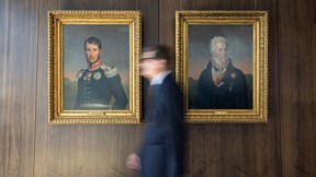 A blurred figure in a dark suit walks past two framed historical portraits hanging on a wood-panelled wall, creating a contrast between motion and tradition in a corporate interior.