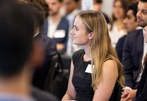 A young woman with blond hair sits at a round table with other peers, watching a training presentation