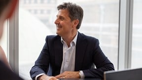 A person wearing a dark suit jacket and a light-coloured collared shirt is seated at a meeting table in a modern office setting.