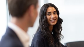 A person with long dark wavy hair is seated in a modern office meeting room, facing another meeting participant who is visible out of focus in the foreground. The person is wearing a dark top and is positioned against a light-coloured background with soft natural lighting. A meeting chair is partially visible in the background. The image uses a shallow depth of field, keeping the seated person in sharp focus while the foreground figure and background remain softly blurred.