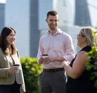 Colleagues standing and talking on an outdoor terrace overlooking the London skyline, with modern office buildings and greenery visible in the background.