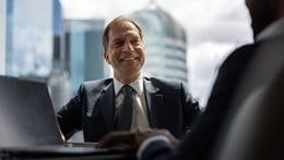 A person wearing a dark suit, white collared shirt and dark tie is seated at a meeting table in a modern office with large floor-to-ceiling windows overlooking a city skyline. An open laptop is positioned on the table to the left, while another meeting participant is visible out of focus in the foreground. Several modern high-rise buildings can be seen through the windows in the background. Natural daylight illuminates the scene, and a shallow depth of field keeps the seated participant in sharp focus while the foreground and cityscape remain softly blurred.