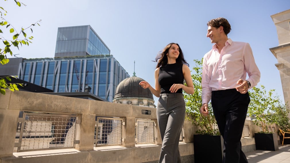 Two colleagues talking on a rooftop terrace overlooking the City of London skyline.