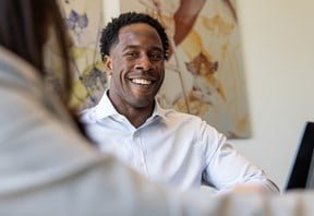 Person in a shirt seated at a desk, speaking with a colleague in an office meeting room.