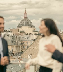 Three people stand on a rooftop overlooking a cityscape. The atmosphere is lively, with one person gesturing animatedly while chatting.