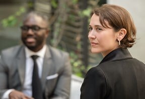 Two people sitting outside, one in a gray suit and tie, smiling, and the other in a black shirt, looking thoughtful. Background shows greenery.