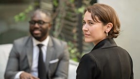 Two people sitting outside, one in a gray suit and tie, smiling, and the other in a black shirt, looking thoughtful. Background shows greenery.