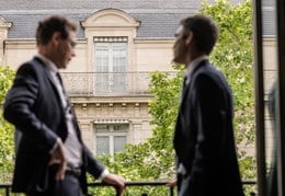 Two people in discussion beside a large window, silhouetted against a historic stone building and leafy trees visible outside.