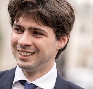 A person in a suit is smiling while engaged in conversation outdoors. The background is blurred, suggesting a casual, pleasant interaction.