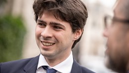 A person in a suit is smiling while engaged in conversation outdoors. The background is blurred, suggesting a casual, pleasant interaction.
