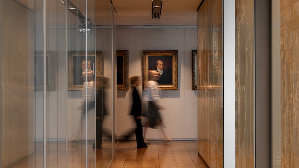 Blurred figures walk through a modern gallery-like cor with a framed portrait painting of NM Rothschild on the far wall.