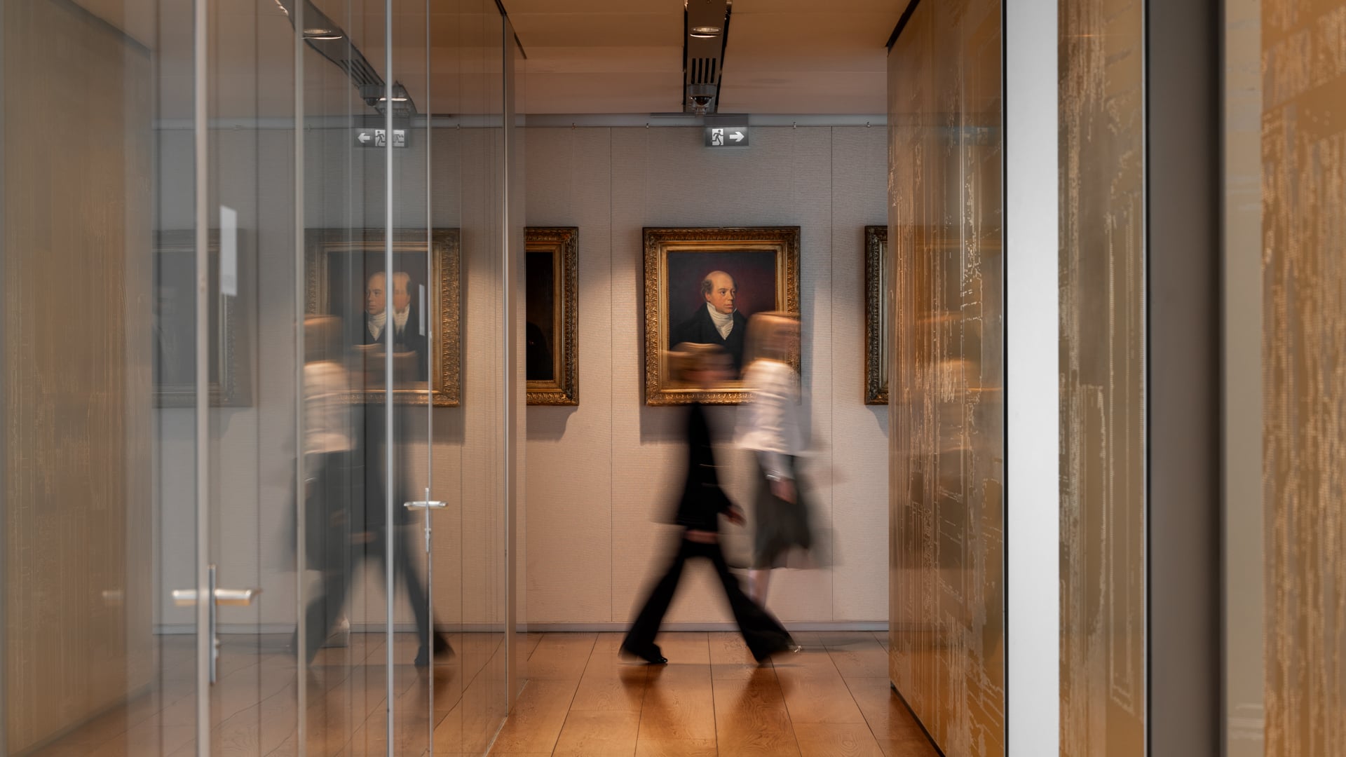 People move through a corridor lined with glass walls and historical portraits, blending modern office life with a sense of tradition.