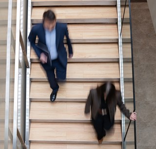 A blurred overhead shot of two people walking on a wooden staircase in a modern building. The movement conveys a sense of urgency and busy office life.