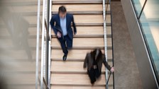 A blurred overhead shot of two people walking on a wooden staircase in a modern building. The movement conveys a sense of urgency and busy office life.