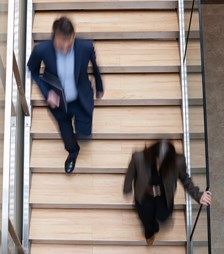 A blurred overhead shot of two people walking on a wooden staircase in a modern building. The movement conveys a sense of urgency and busy office life.