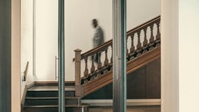 A person stands on an indoor staircase, seen through a large window. The scene is calm, with soft light creating a peaceful ambiance.