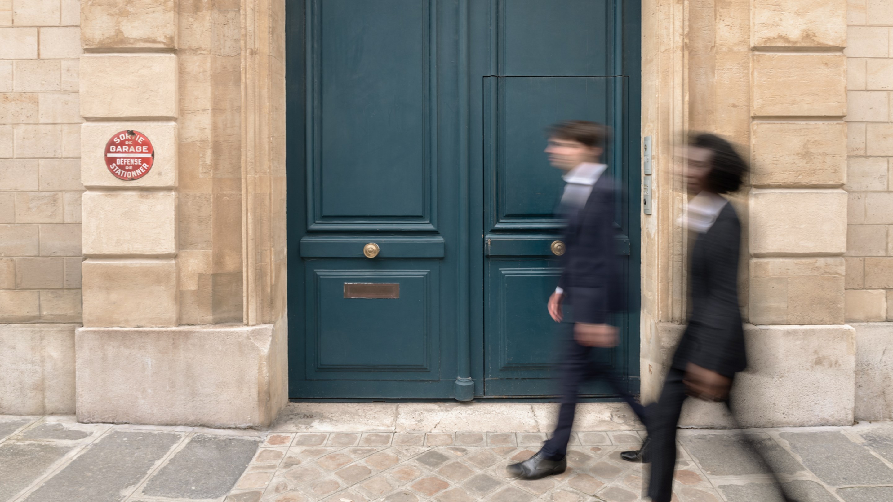 Two people walk past a large blue door set in a stone wall. They both wear dark clothing and appear engaged in conversation on a cobblestone street.