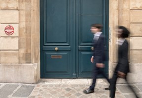 Two people walk past a large blue door set in a stone wall. They both wear dark clothing and appear engaged in conversation on a cobblestone street.