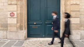 Two people walk past a large blue door set in a stone wall. They both wear dark clothing and appear engaged in conversation on a cobblestone street.