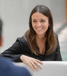 Colleague seated at a table, speaking with another colleague in a professional office environment.
