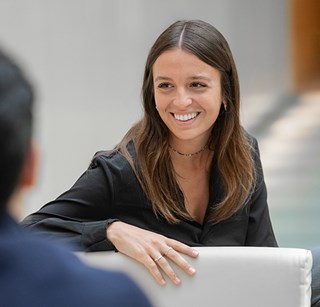 Colleague seated at a table, speaking with another colleague in a professional office environment.