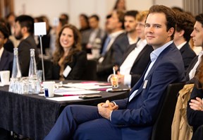 Attendees seated at round tables during a training event, listening to a speaker. A participant in a blue suit is in focus in the foreground, with notebooks, glasses and water bottles visible on the tables.