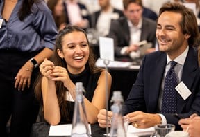 Attendees seated together at a training event. Two participants in the foreground are engaged in conversation at a table with notepads and water bottles.