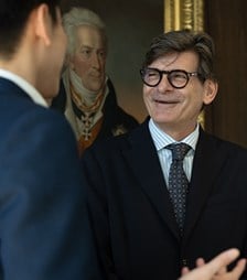 Two colleagues in business attire conversing in an office environment, with one colleague gesturing while speaking.