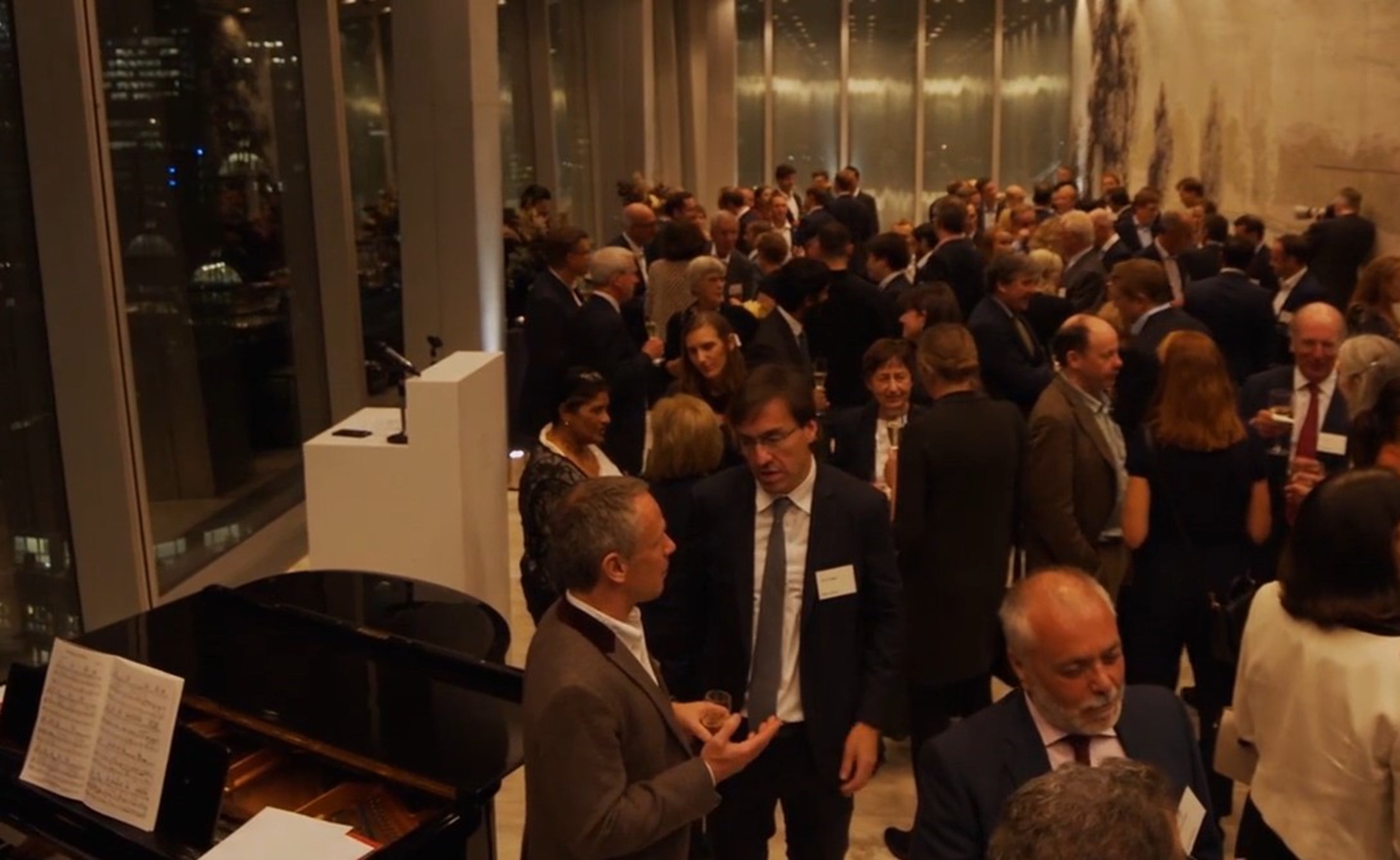 Attendees gather at a Rothschild & Co Alumni Network networking reception in a high-rise venue at night, with floor-to-ceiling windows overlooking a city skyline. Groups of guests converse throughout the room, while a grand piano and a lectern stand in the foreground. Warm lighting and large wall artwork create a formal event atmosphere.
