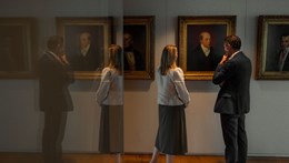 Two visitors viewing framed portraits in a gallery with reflections visible in the foreground