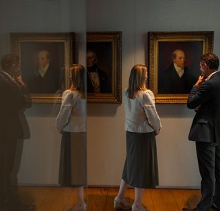 Two visitors viewing framed portraits in a gallery with reflections visible in the foreground
