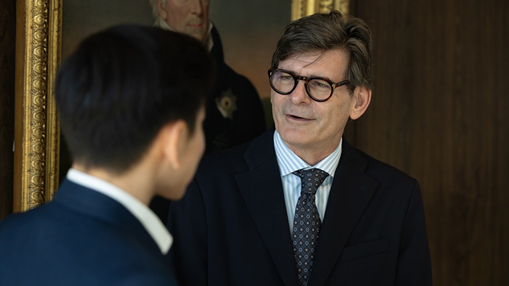 Image of a man having a conversation with someone, stood in front of an oil painting of one of the Rothschild family