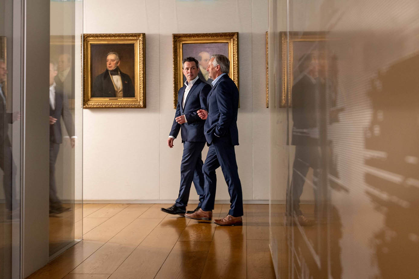 Two men walking down a hallway behind family paintings