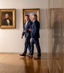 Two men walking down a hallway behind family paintings