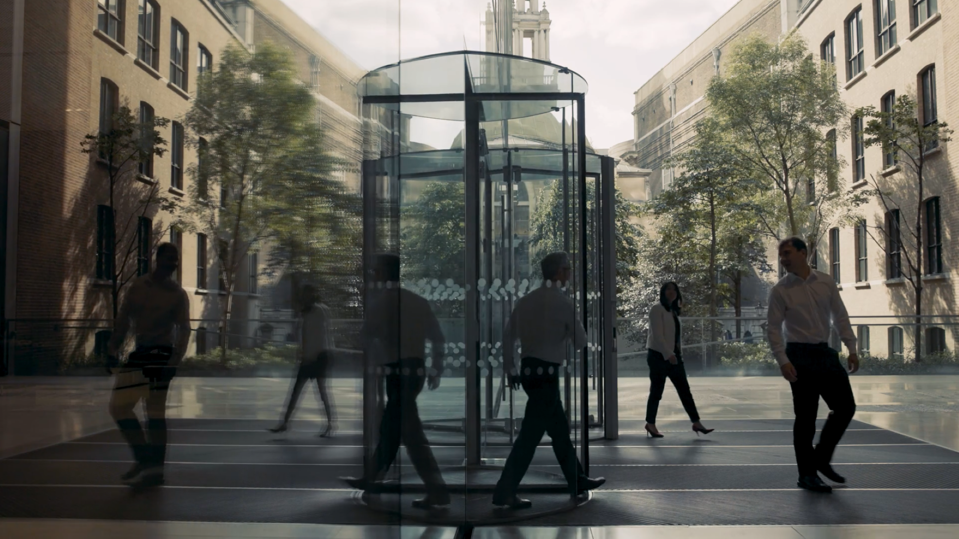 People move through a glass revolving door in a modern office entrance, with reflections, motion blur and tree-lined courtyards visible through the floor-to-ceiling windows.