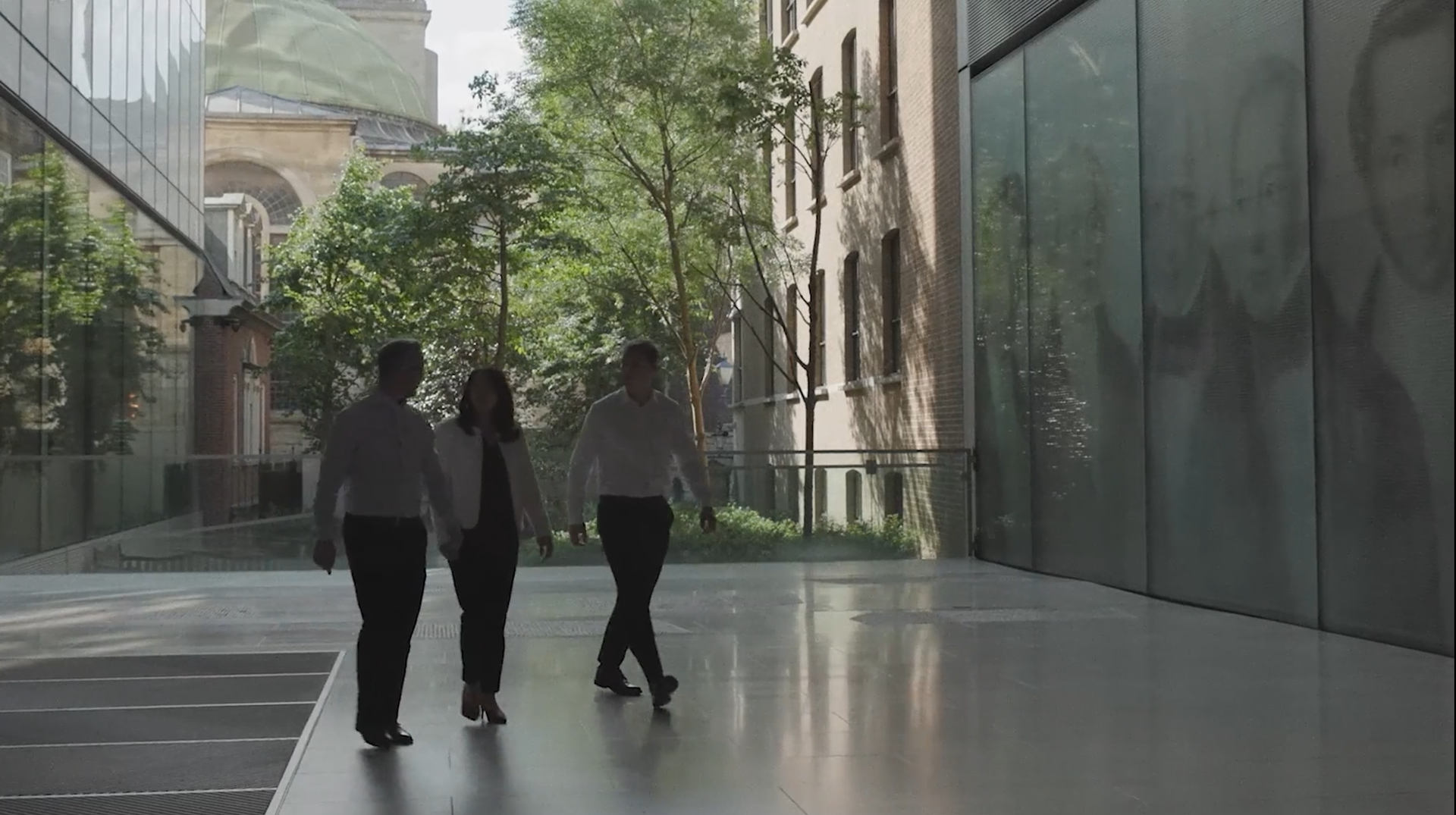 Three people walk through a bright, modern courtyard between glass and stone buildings, with trees and a domed historic building visible in the background.