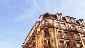 An image of the Parisian office from the street corner, showing only the top half of the stone building with blue skies behind.