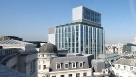 A modern photograph of New Court taken from the same level of another building. The modern glass 16-storey building is surrounded by lower level London typical brick buildings. The background is a blue sky with some airplane lines running through.